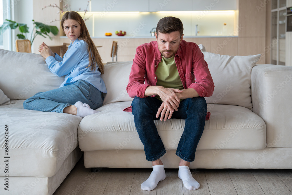 Gloomy upset passive man sitting on sofa apathetically looking on floor with blank stare, and offended woman with reproaching face expression. Misunderstanding, confrontation, crisis in family life