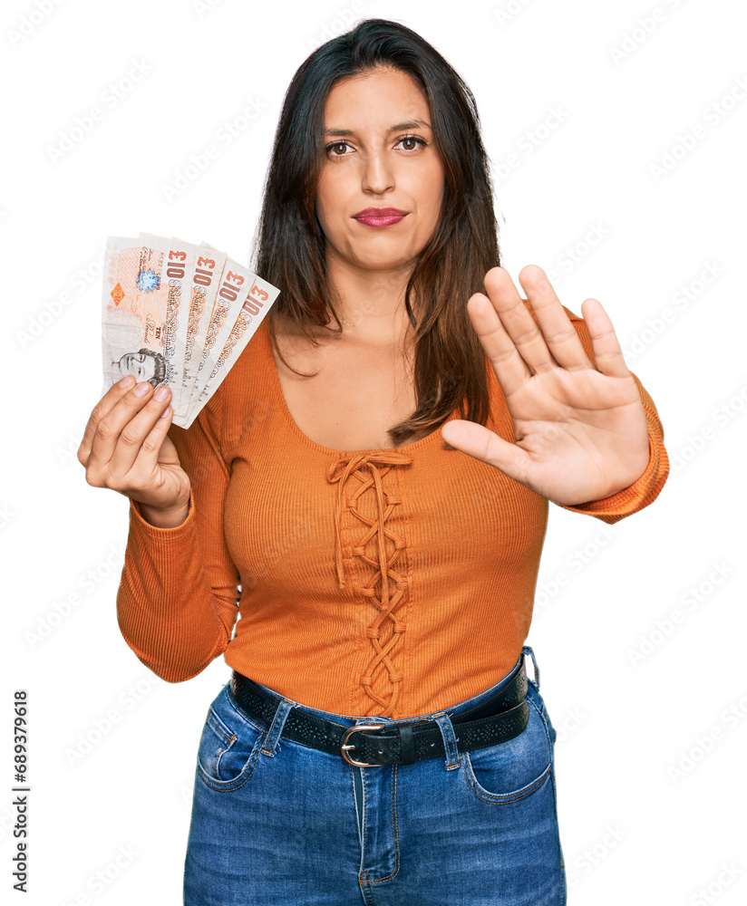 Beautiful hispanic woman holding 10 united kingdom pounds banknotes with open hand doing stop sign with serious and confident expression, defense gesture Beautiful hispanic woman holding 10 united kingdom pounds banknotes with open hand doing stop sign with serious and confident expression, defense gesture