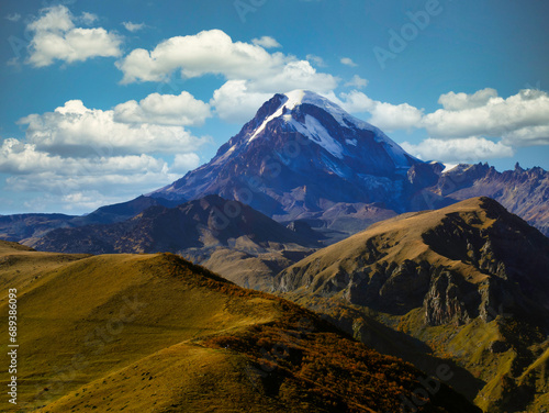 Majestic mountain peak rising above rolling hills under a vivid blue sky dotted with clouds. The contrast of the rugged mountain summit against the soft curves of the surrounding landscape highlights 