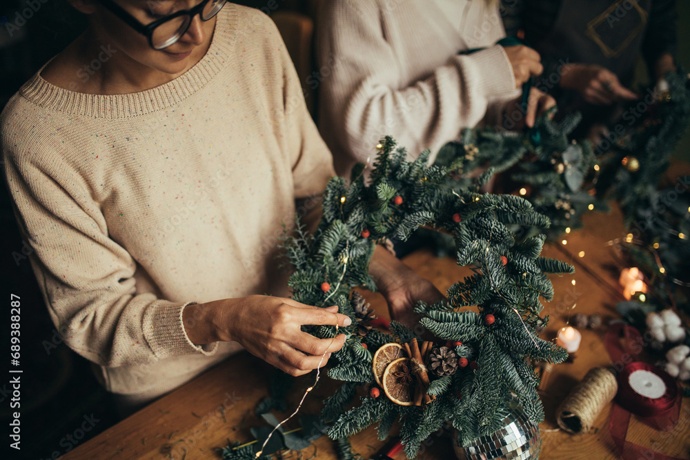 Happy women making Christmas wreath using natural pine branches and festive decorations.
