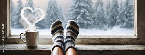 Cozy feet in wool socks by a windowsill, with a steaming cup and a heart-shaped steam above, overlooking a snowy forest. Panorama with copy space. White background.