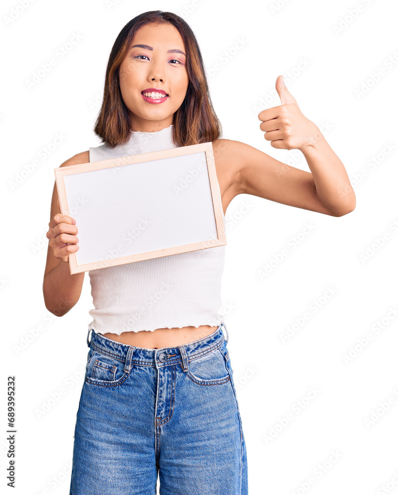 Young beautiful chinese girl holding empty white chalkboard smiling happy and positive, thumb up doing excellent and approval sign Young beautiful chinese girl holding empty white chalkboard smiling happy and positive, thumb up doing excellent and approval sign
