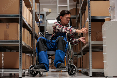 Warehouse asian package handler in wheelchair searching cardboard box on shelf. Shipment manager with disability taking parcel from rack while working in inclusive workplace photo