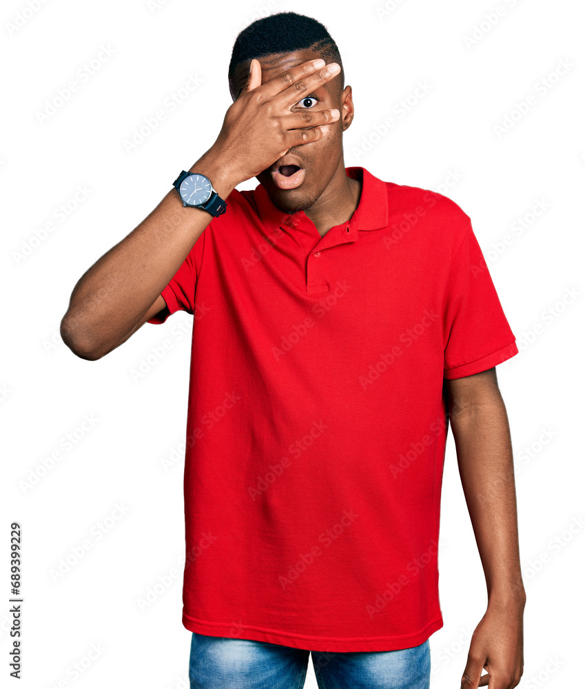 Young african american man wearing casual red t shirt peeking in shock covering face and eyes with hand, looking through fingers with embarrassed expression. Young african american man wearing casual red t shirt peeking in shock covering face and eyes with hand, looking through fingers with embarrassed expression.