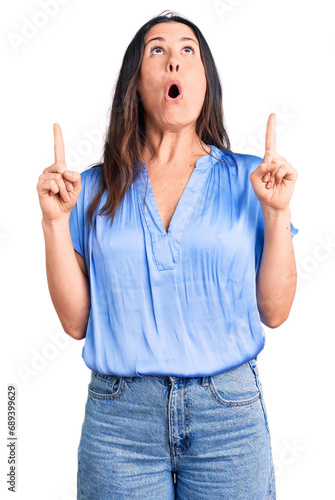 Young beautiful brunette woman wearing casual t-shirt amazed and surprised looking up and pointing with fingers and raised arms.