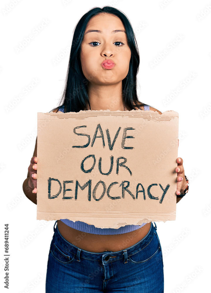 Beautiful hispanic woman holding save our democracy protest banner puffing cheeks with funny face. mouth inflated with air, catching air. Beautiful hispanic woman holding save our democracy protest banner puffing cheeks with funny face. mouth inflated with air, catching air.