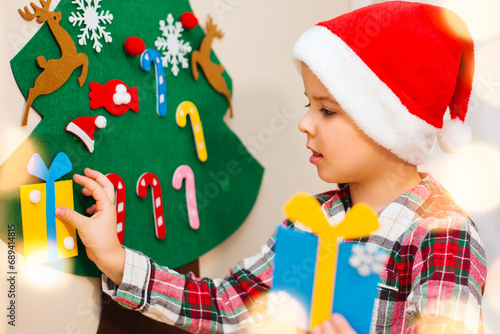 A cute seven-year-old boy in red plaid pajamas and a Santa hat decorates his felt hand-made Christmas tree, which hangs on the wall at home.