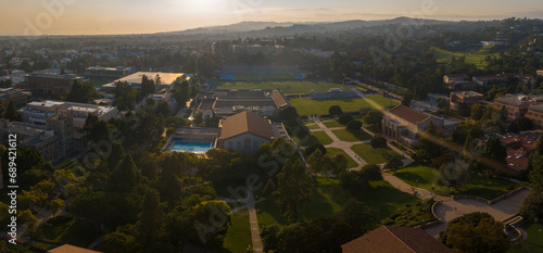 Aerial sunset view of UCLA campus, showcasing a blend of classic and modern architecture amid verdant foliage, sports facilities, and a tranquil backdrop of hills and water.