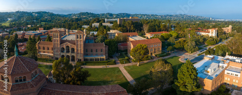 Aerial view of UCLA campus with Gothic tower, red-brick buildings, green lawns, and pathways amidst hilly, tree-covered landscape in soft, golden light.