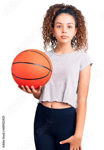 Beautiful kid girl with curly hair holding basketball ball thinking attitude and sober expression looking self confident