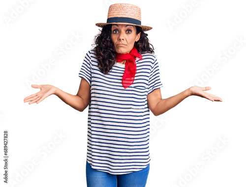 Middle age beautiful woman wearing casual striped t shirt and summer hat clueless and confused expression with arms and hands raised. doubt concept.