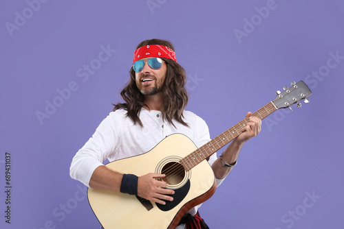 Stylish hippie man in sunglasses playing guitar on violet background