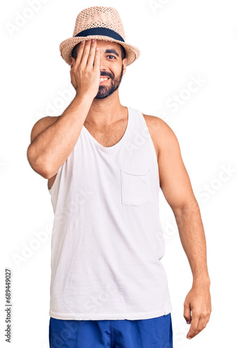 Young hispanic man wearing casual summer hat covering one eye with hand, confident smile on face and surprise emotion.