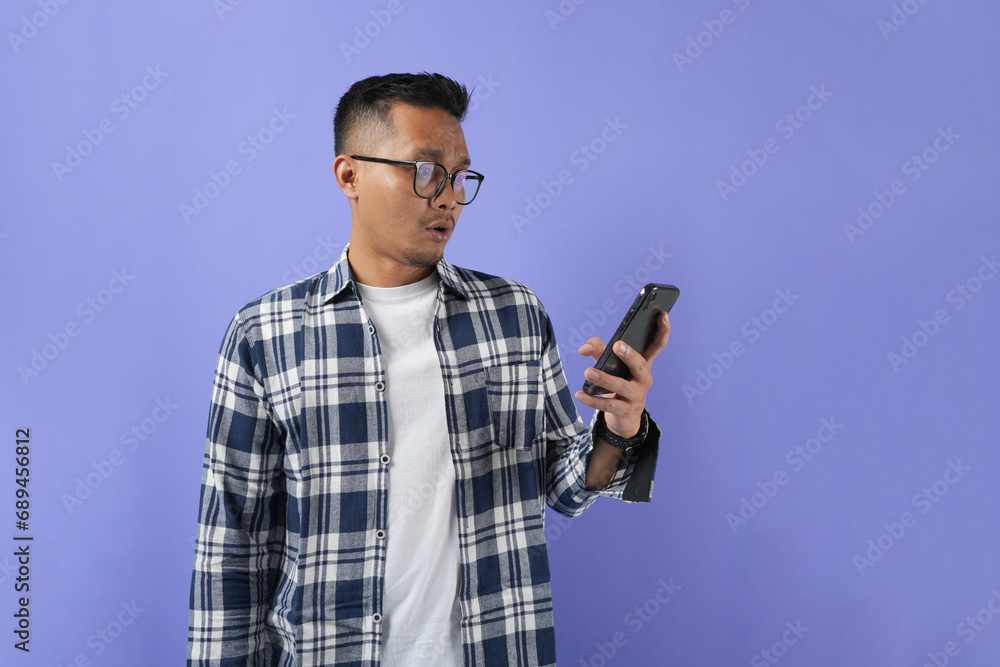 Surprised young Asian man using a mobile phone with a positive expression, wearing a casual shirt, and standing isolated on a purple background. Surprised young Asian man using a mobile phone with a positive expression, wearing a casual shirt, and standing isolated on a purple background.