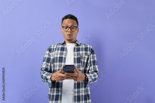 Surprised young Asian man using a mobile phone with a positive expression, wearing a casual shirt, and standing isolated on a purple background. 