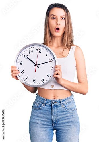 Young beautiful girl holding big clock scared and amazed with open mouth for surprise, disbelief face