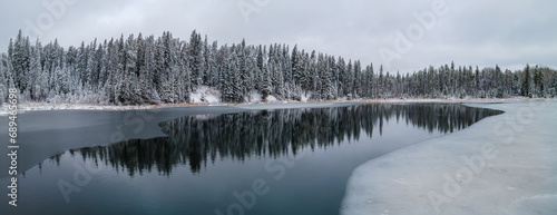 A panoramic view of a cold looking winter scene at a partially frozen lake that is surrounded by an evergreen forest. The trees are covered with heavy snow. Tree reflections can be seen in the calm w