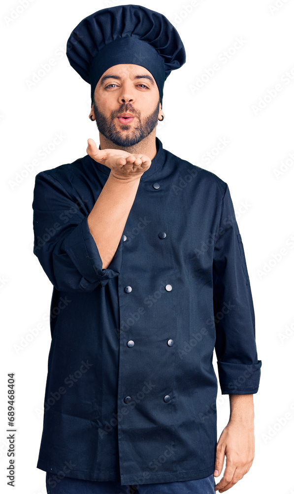 Young hispanic man wearing cooker uniform looking at the camera blowing a kiss with hand on air being lovely and sexy. love expression. Young hispanic man wearing cooker uniform looking at the camera blowing a kiss with hand on air being lovely and sexy. love expression.