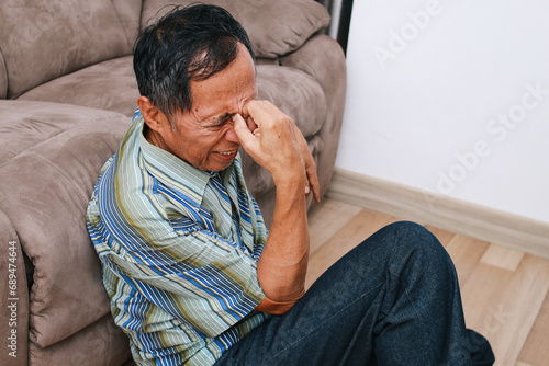 Old senior man sitting on the floor in living room touching forehead suffering from severe headache