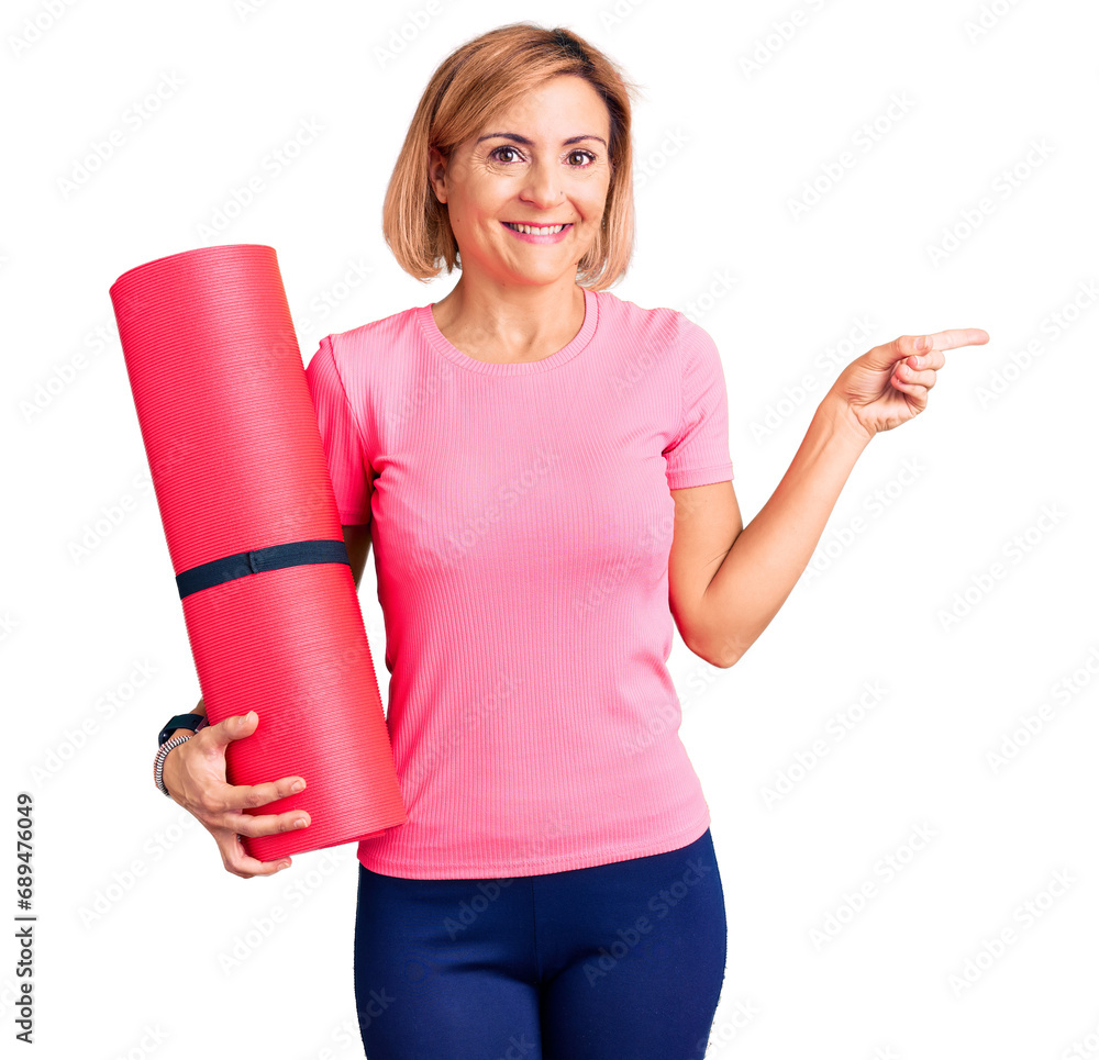 Young blonde woman holding yoga mat smiling happy pointing with hand and finger to the side Young blonde woman holding yoga mat smiling happy pointing with hand and finger to the side