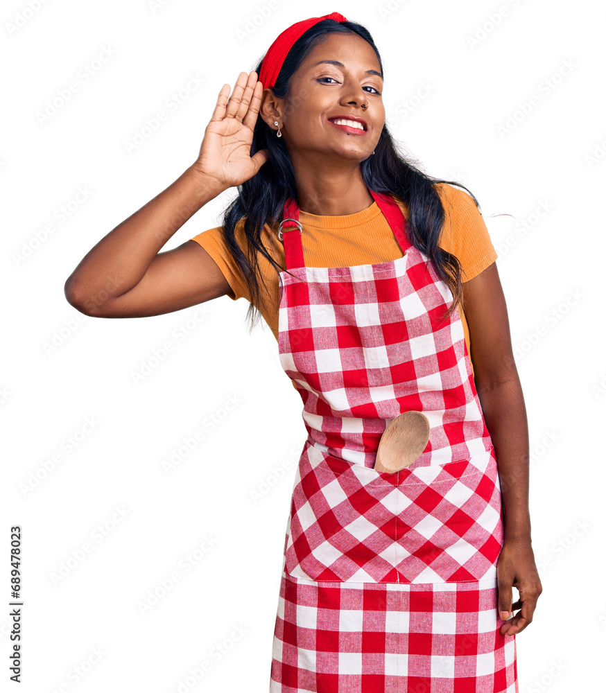 Young indian girl wearing professional baker apron smiling with hand over ear listening an hearing to rumor or gossip. deafness concept. Young indian girl wearing professional baker apron smiling with hand over ear listening an hearing to rumor or gossip. deafness concept.