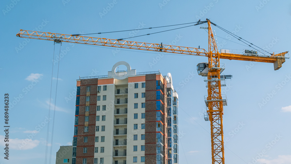 A tower construction crane on the background of a blue sky with clouds. Boom rotary crane with boom. Construction of apartment buildings in the city. Tower crane cabin. A tower construction crane on the background of a blue sky with clouds. Boom rotary crane with boom. Construction of apartment buildings in the city. Tower crane cabin.