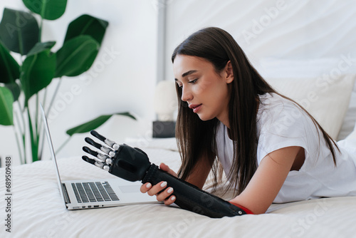 Young girl attaching artificial cyber arm laying on bed using laptop exploring instructions of electronic bionic limb. Hi tech medicine, future technologies. photo