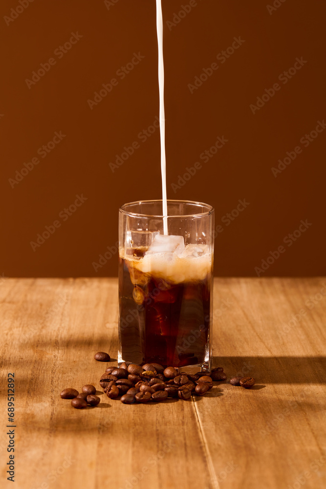 A stream of milk is pouring from above into a cup of black coffee on a wooden table with a brown background. Art space for food and beverage advertising. A stream of milk is pouring from above into a cup of black coffee on a wooden table with a brown background. Art space for food and beverage advertising.
