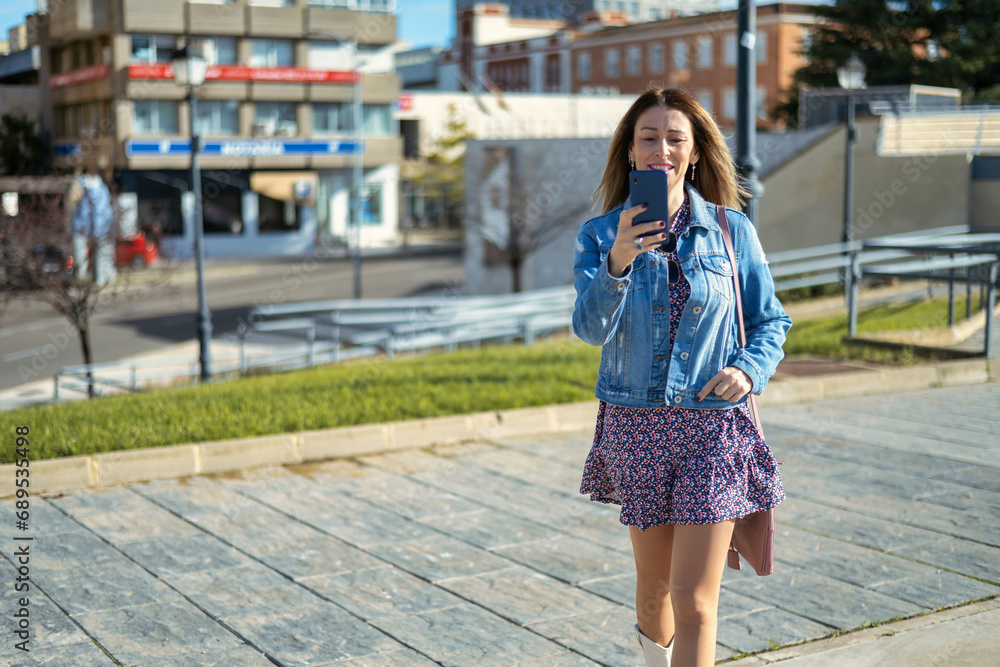 Naklejka premium Young Woman Reading A Message On The Phone While Walking In The City