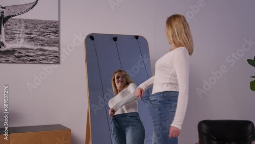 Cheerful charming mature woman in too big jeans looking at reflection in mirror, demonstrating and enjoying weight loss diet achievement, expressing excitement and happiness in domestic room. photo