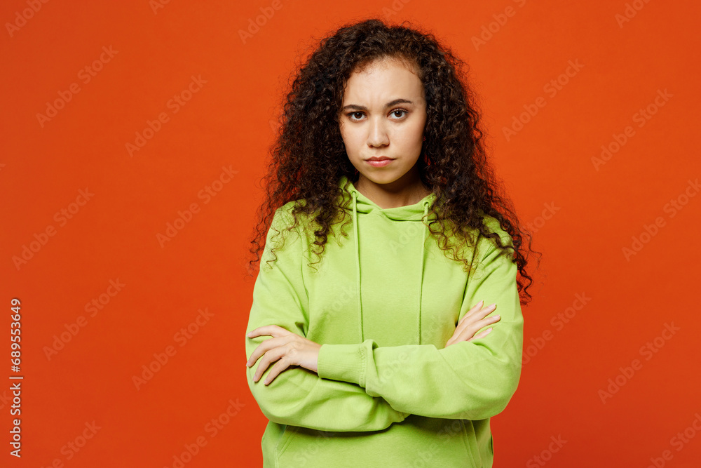 Young sad frowning grumpy woman of African American ethnicity she wearing green hoody casual clothes hold hands crossed folded look camera isolated on plain red orange background. Lifestyle concept. Young sad frowning grumpy woman of African American ethnicity she wearing green hoody casual clothes hold hands crossed folded look camera isolated on plain red orange background. Lifestyle concept.