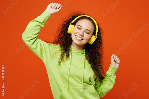Young cheerful fun cool smiling woman of African American ethnicity she wear green hoody casual clothes listen to music in headphones dance isolated on plain red orange background. Lifestyle concept. photo