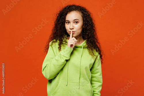 Young secret fun woman of African American ethnicity she wear green hoody casual clothes say hush be quiet with finger on lips shhh gesture isolated on plain red orange background. Lifestyle concept.
