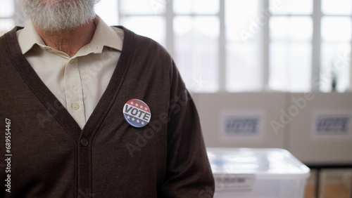 A close-up of a senior man with a vote button on his chest standing in the hall during the elections in the US photo