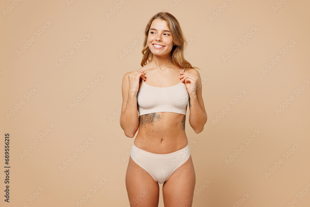 Fototapeta premium Young smiling happy nice lady woman with slim body perfect skin wear nude lingerie stand hold top bra breast looking aside isolated on plain pastel light beige background. Lifestyle diet fit concept.