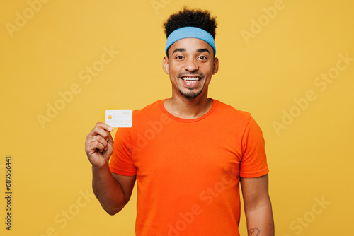 Young fitness trainer instructor sporty man sportsman wears orange t-shirt hold mock up of credit bank card spend time in home gym isolated on plain yellow background. Workout sport fit abs concept.