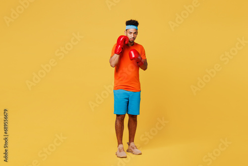 Full body young fitness trainer instructor sporty man sportsman wear orange t-shirt boxing red gloves punch spend time in home gym isolated on plain yellow background. Workout sport fit abs concept.