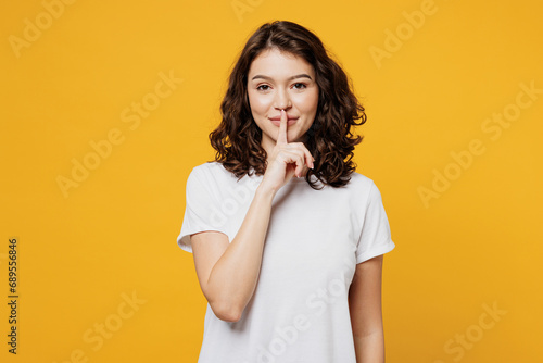 Young secret smiling Caucasian woman she wearing white blank t-shirt casual clothes saying hush be quiet with finger on lips shhh gesture isolated on plain yellow orange background. Lifestyle concept. photo