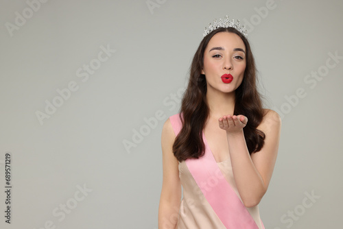 Beautiful young woman with tiara and ribbon in dress blowing kiss on grey background, space for text. Beauty contest