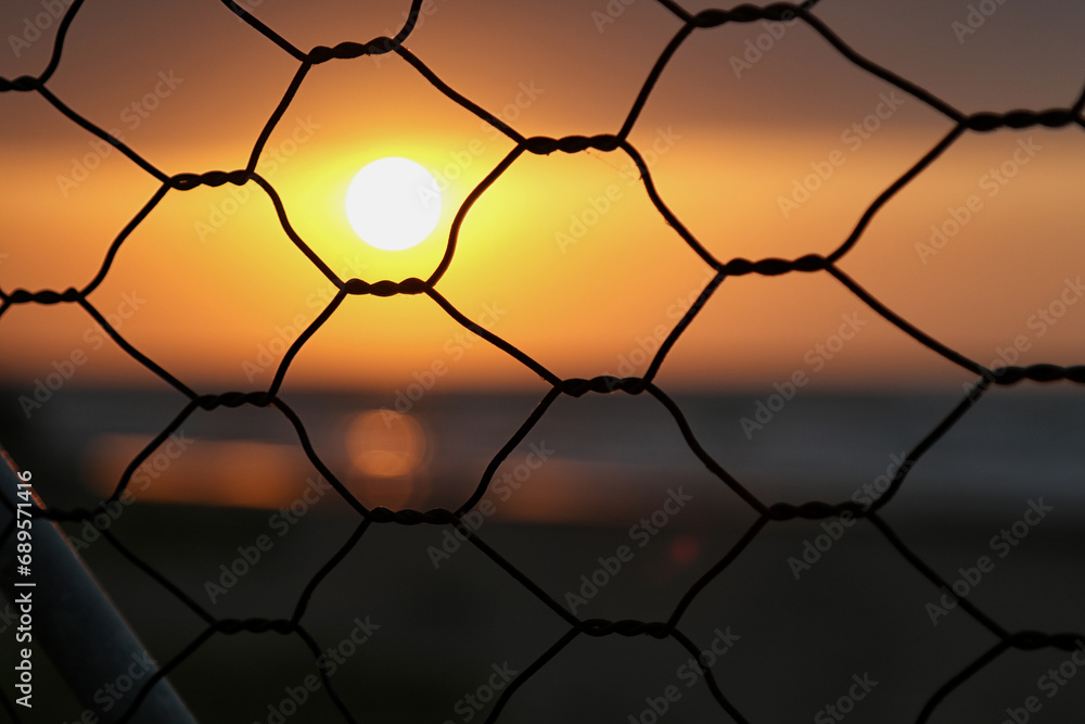 Fototapeta premium The sunset's orange glow reflecting off the sea, seen through a fence adorned with a spider's web.