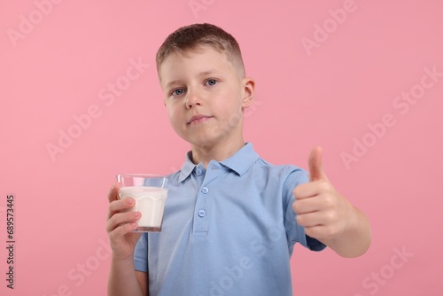 Cute boy with glass of fresh milk showing thumb up on pink background