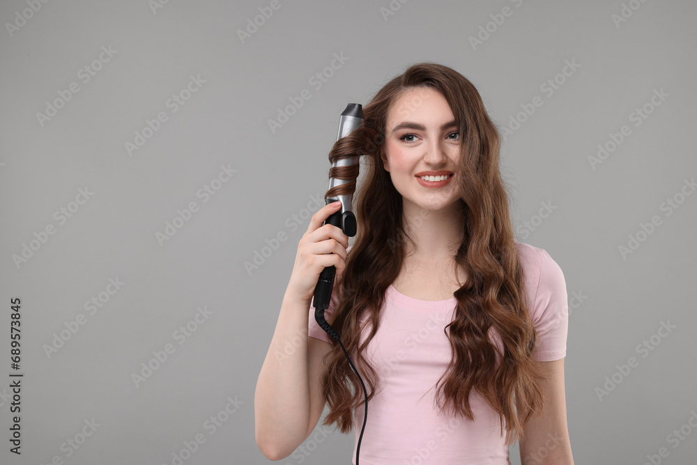 Beautiful young woman using curling hair iron on grey background, space for text Beautiful young woman using curling hair iron on grey background, space for text