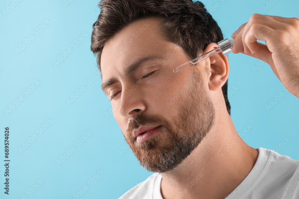 Obraz premium Handsome man applying cosmetic serum onto his face on light blue background, closeup. Space for text