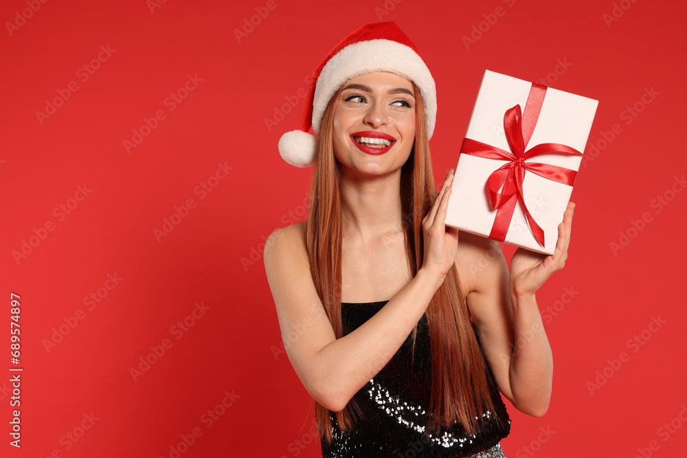 Young woman in Santa hat with Christmas gift on red background, space for text Young woman in Santa hat with Christmas gift on red background, space for text