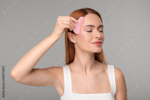 Young woman massaging her face with rose quartz gua sha tool on grey background photo