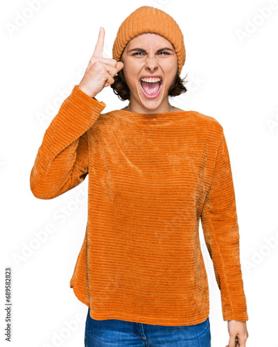 Young hispanic woman wearing casual clothes and wool cap pointing finger up with successful idea. exited and happy. number one.