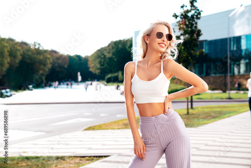 Fototapeta Naklejka Na Ścianę i Meble -  Young beautiful smiling hipster woman in trendy summer white top and violet pants clothes. Carefree model posing in the street at sunset. Positive blond female. Cheerful and happy. In sunglasses