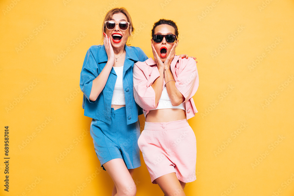 Two young beautiful smiling brunette hipster female in trendy summer clothes. Sexy carefree women near yellow wall in studio. Positive models having fun. Cheerful and happy. Shocked and surprised - obrazy, fototapety, plakaty 