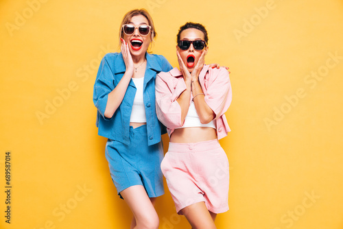Two young beautiful smiling brunette hipster female in trendy summer clothes. Sexy carefree women near yellow wall in studio. Positive models having fun. Cheerful and happy. Shocked and surprised