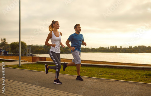 Happy smiling sporty couple wearing sportswear jogging along the river in the park having sport workout. Young man and woman running outdoors. Workout in nature and healthy lifestyle concept.